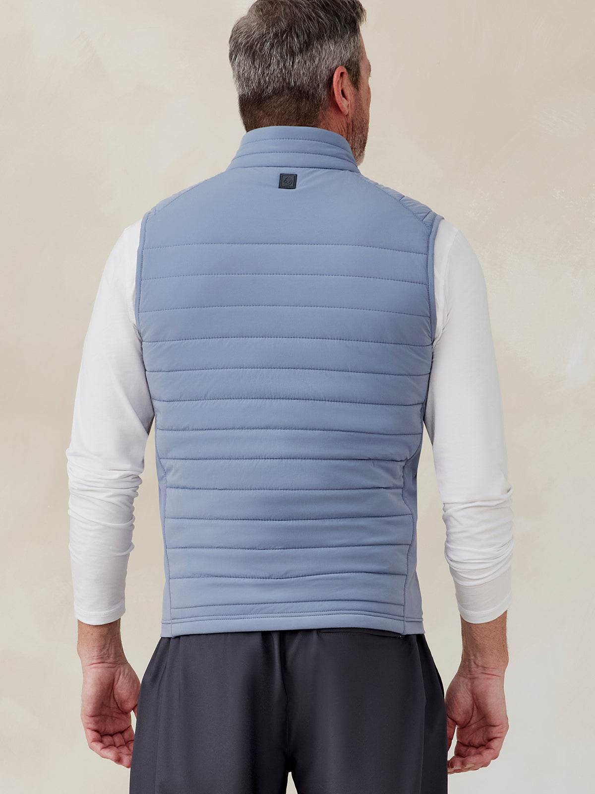 Man wearing light blue quilted hybrid vest with stand collar, shown from back, paired with white long sleeve shirt and dark pants tasc Hilltop Hybrid Vest (Anchor)