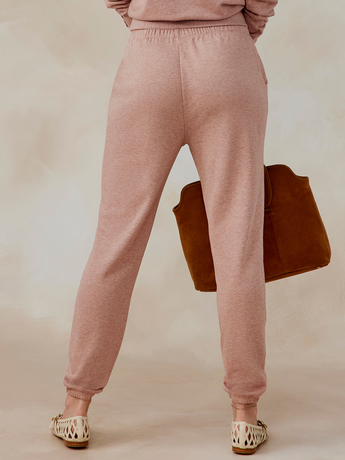 Back view of dusty pink Stadium French Terry sweatpants with elastic waistband and cuffs, worn by model holding brown bag tasc performance stadium sweatpant (AntiqueRosyHeather)