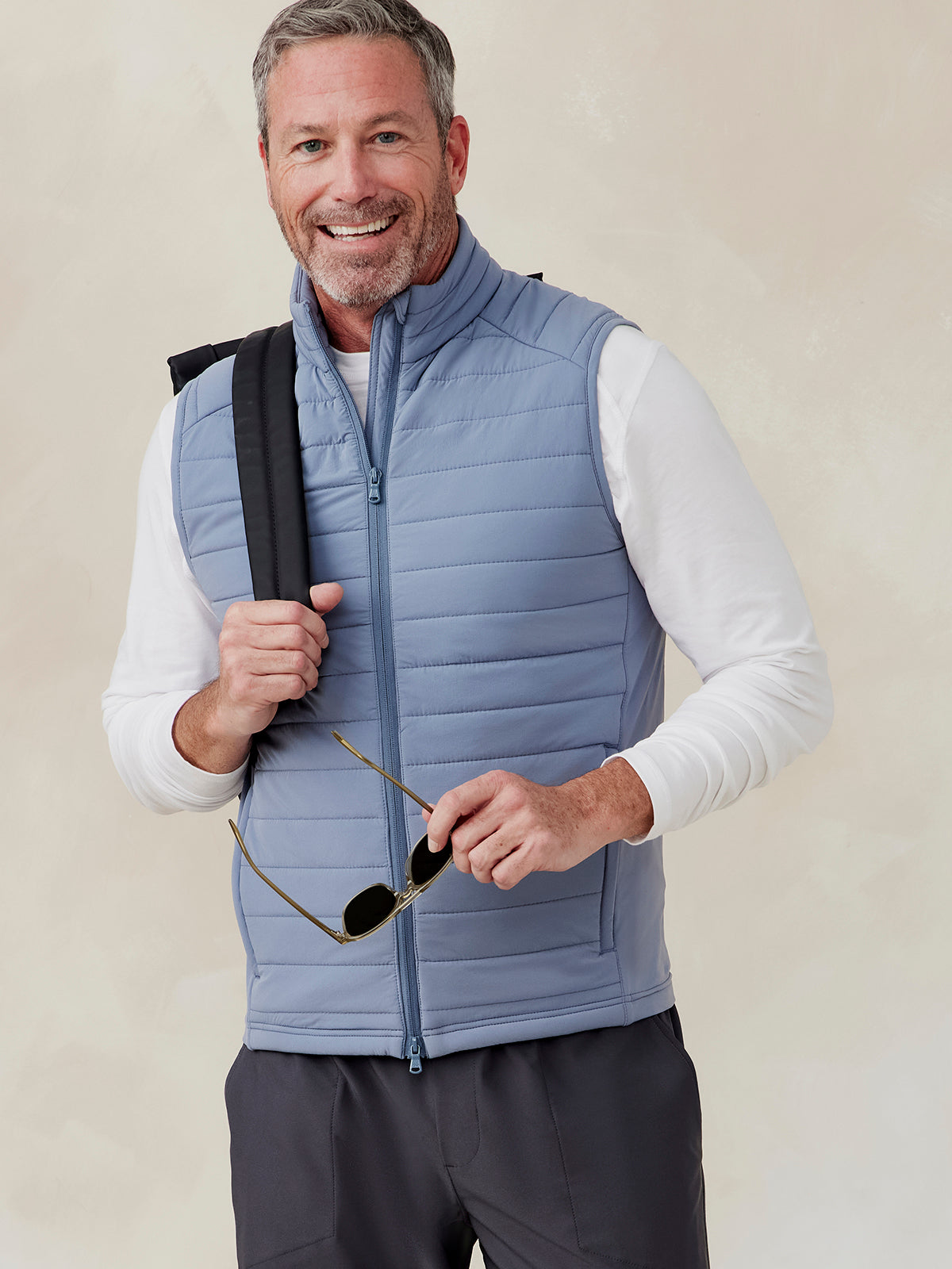Man wearing a light blue quilted Hilltop Hybrid vest with a front zipper over a white long-sleeve shirt, holding sunglasses tasc Hilltop Hybrid Vest (Anchor)