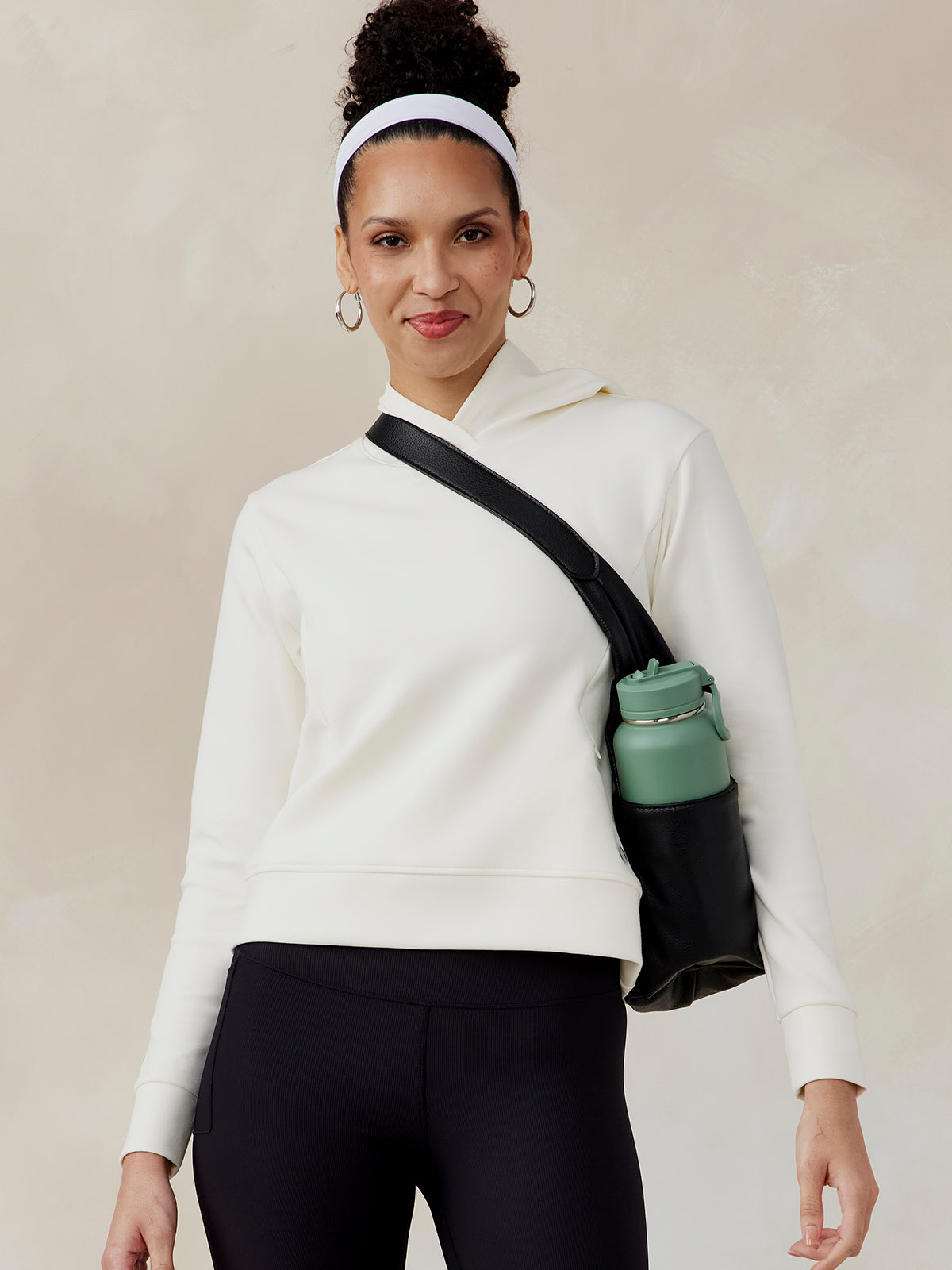 Woman wearing a white Highlands Hoodie with a hood, paired with black pants and a black crossbody bag holding a green water bottle tasc Highlands Hoodie (Coconut)
