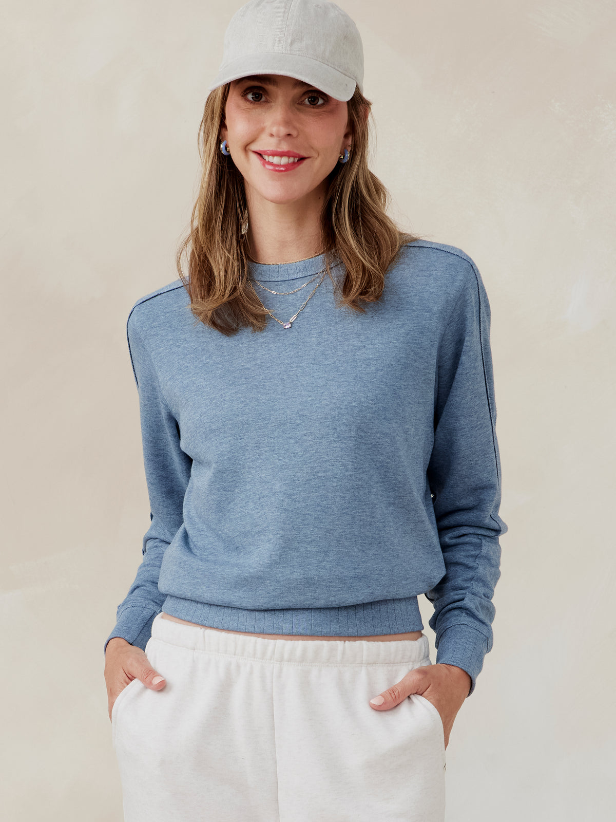 Woman wearing a blue French terry sweatshirt with ribbed hem and cuffs, paired with white pants and a light gray cap tasc Stadium Sweatshirt (AnchorHeather)