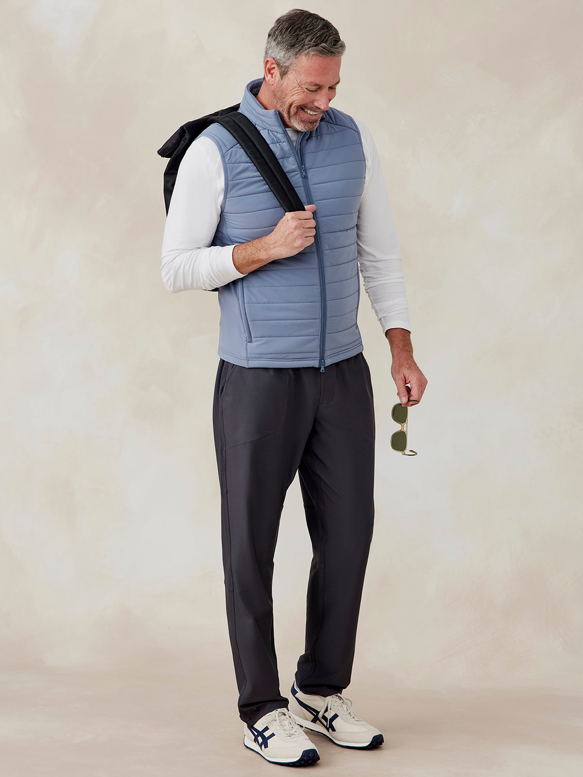 Man wearing a light blue quilted zip-up vest over a white long-sleeve shirt, dark pants, and white sneakers, holding sunglasses tasc Hilltop Hybrid Vest (Anchor)
