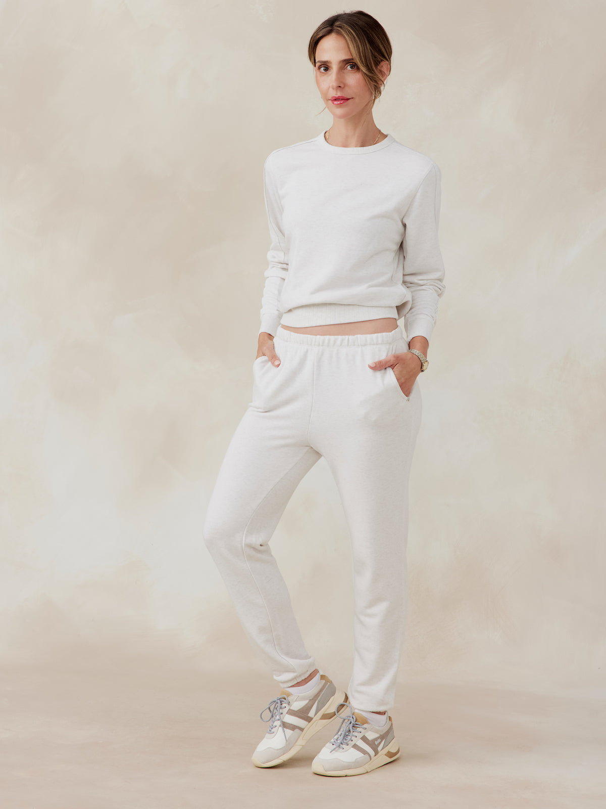 Woman wearing light beige Stadium French Terry sweatshirt and matching jogger pants with sneakers, front view, hands in pockets tasc Stadium Sweatshirt (CoconutHeather)