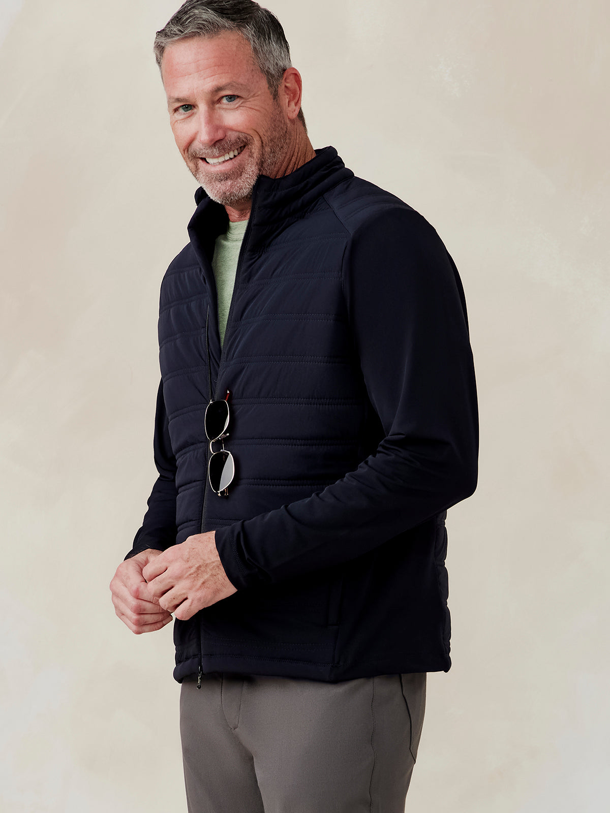 Man wearing a black Hilltop Hybrid Jacket with quilted front and smooth sleeves, zipped halfway, holding sunglasses Hilltop Hybrid Jacket (Black)
