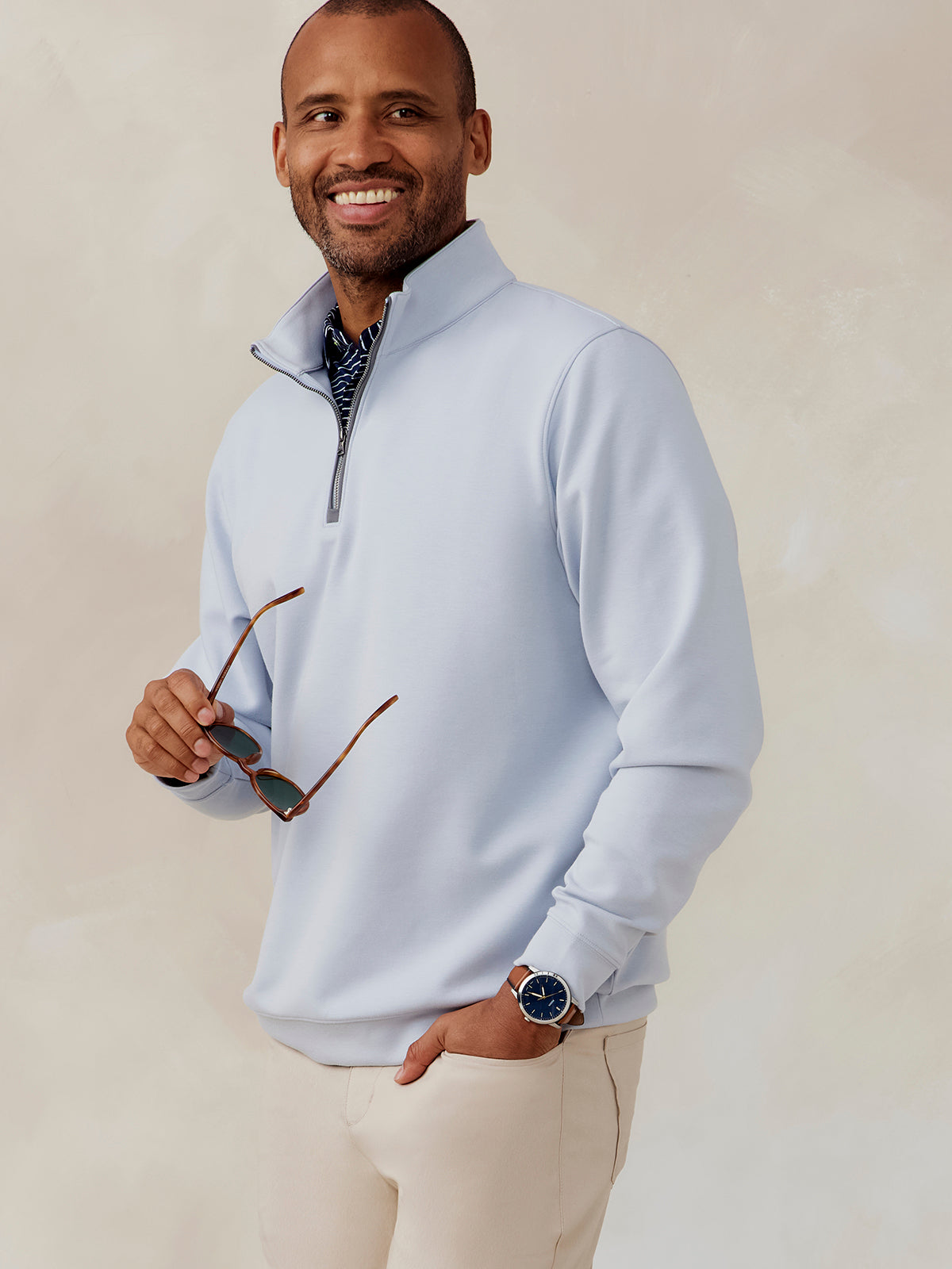Man wearing light blue Highlands quarter zip pullover with beige pants, holding sunglasses and wearing a watch with a blue face tasc Highlands Quarter Zip (Cloud)