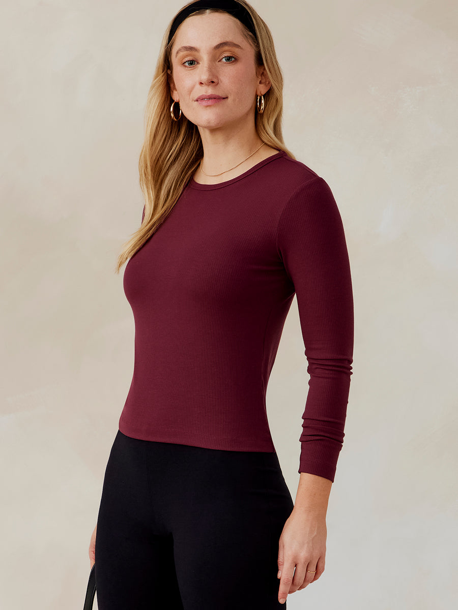 Woman wearing a fitted long sleeve ribbed top in dark burgundy, front and side view, paired with black pants tasc Microluxe Rib LS (Wineberry)