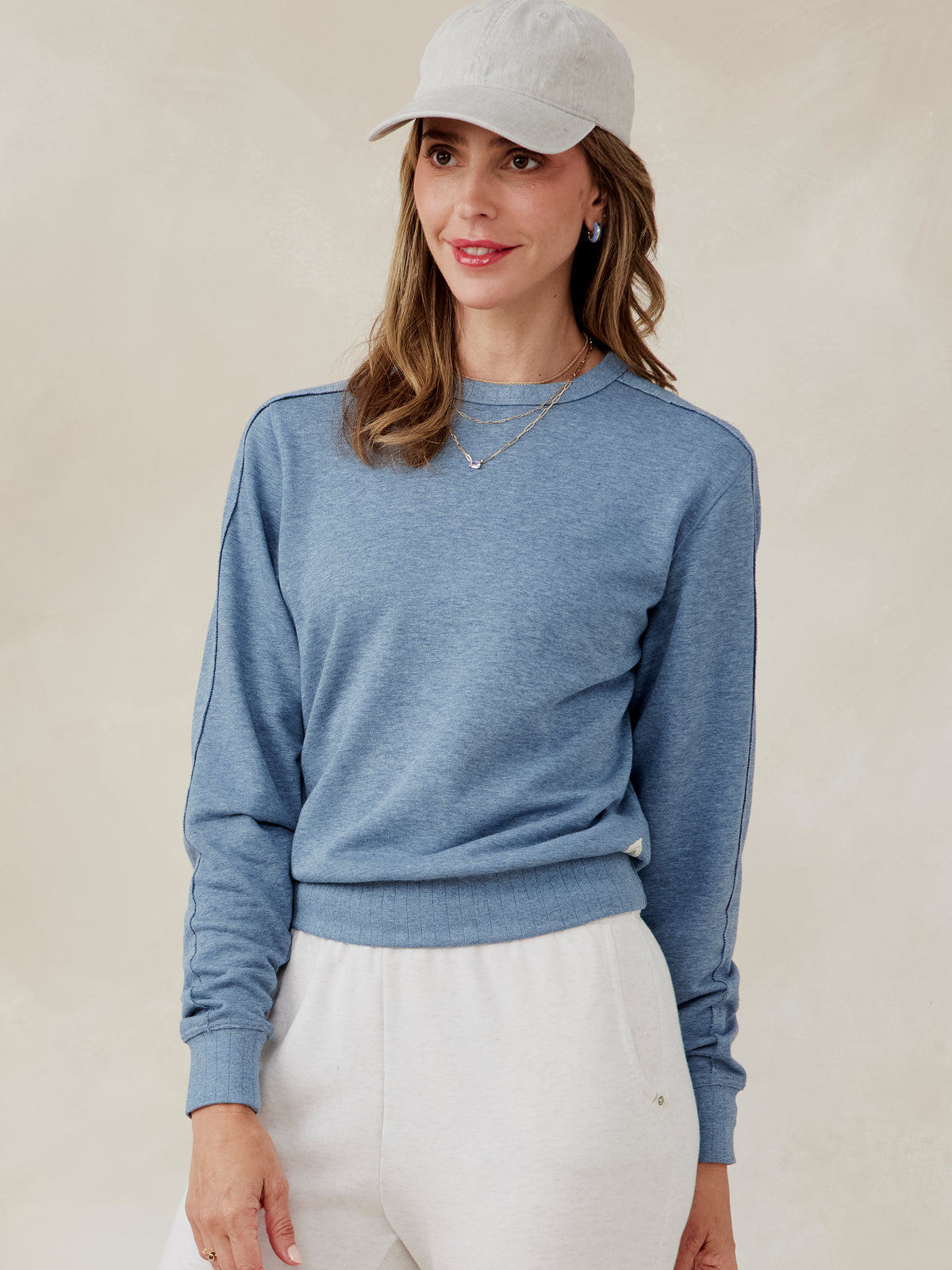 Woman wearing a blue French terry sweatshirt with ribbed cuffs and hem, paired with white pants and a light gray cap tasc Stadium Sweatshirt (AnchorHeather)